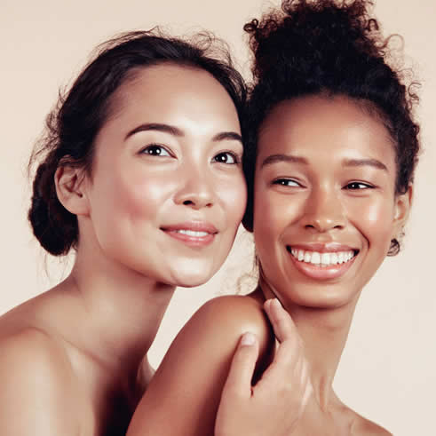 Beauty Image of two women with glowing skin smiling while showing off their clean makeup looks.