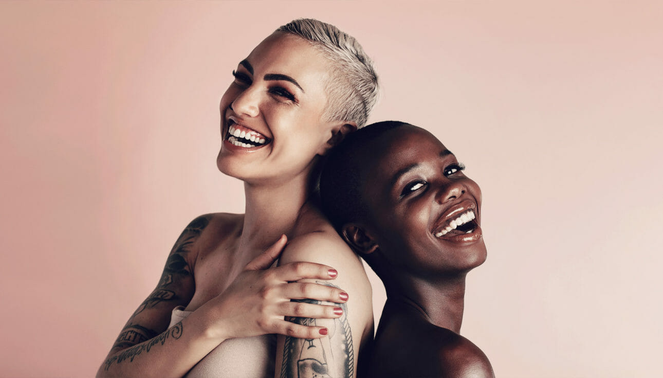 Two women smiling with clean beauty products on their faces on a peach background.
