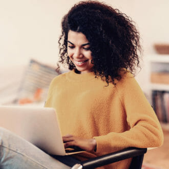 Woman in a yellow sweater smiling at her laptop while shopping for clothing.