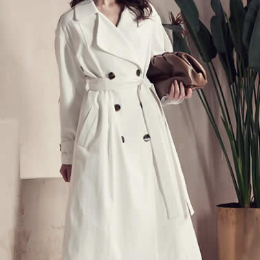 Female fashion model posing in a white belted peacoat near a potted plant. Fashion Product Attribution.