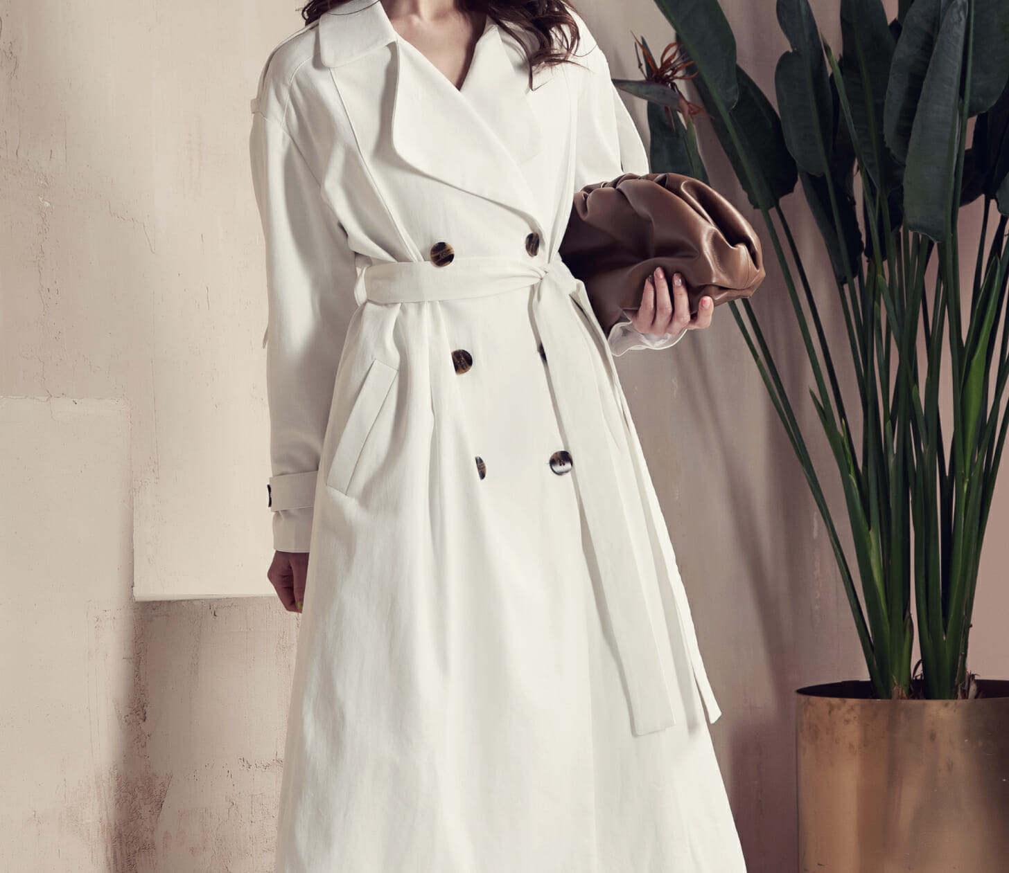 Female fashion model posing in a white belted peacoat near a potted plant.